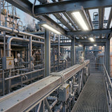 Industrial plant interior featuring a metal walkway, intricate piping, pressure gauges, and bright linear high-bay LED lights.